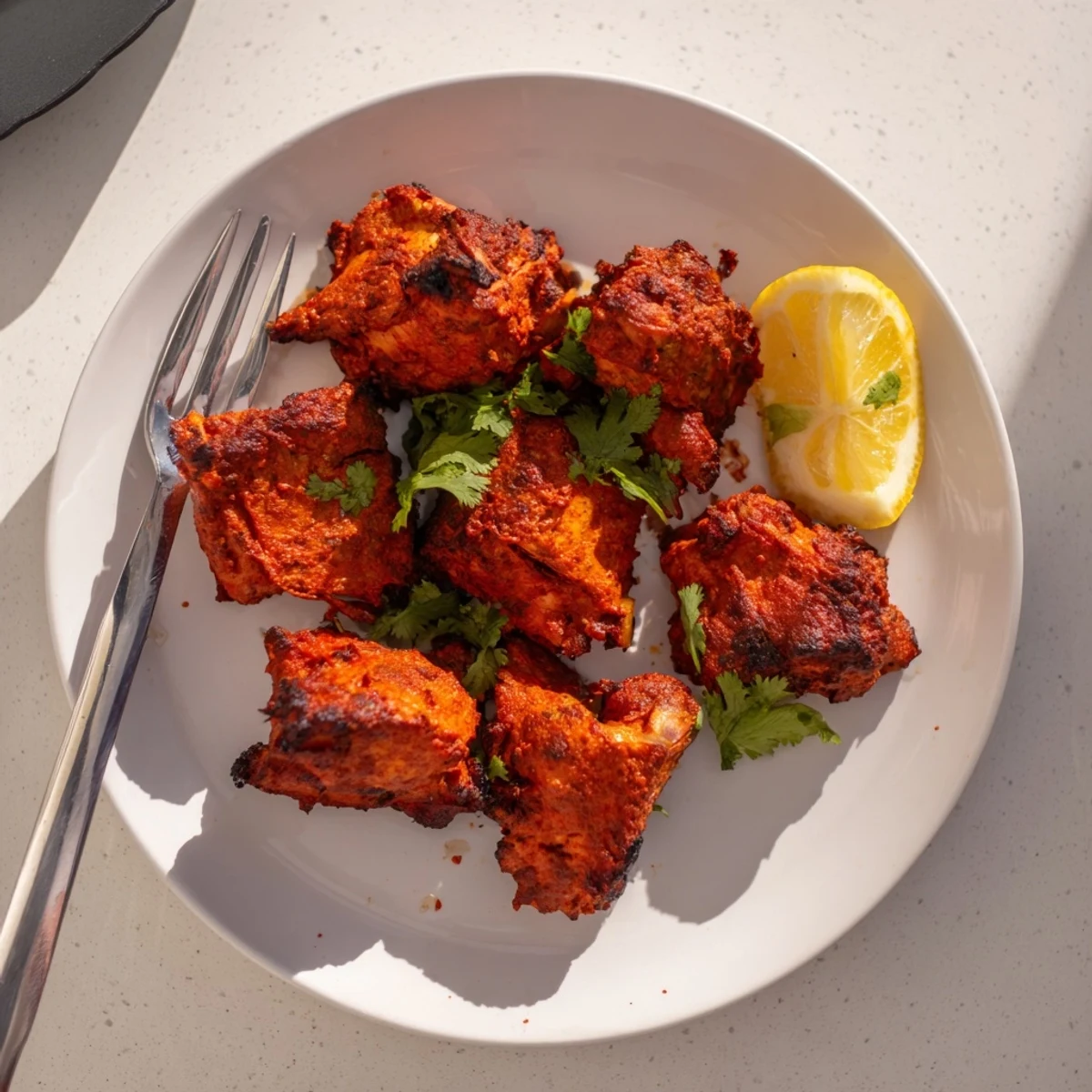 Tandoori chicken with charred edges served with fresh cilantro and lemon wedges on a white plate